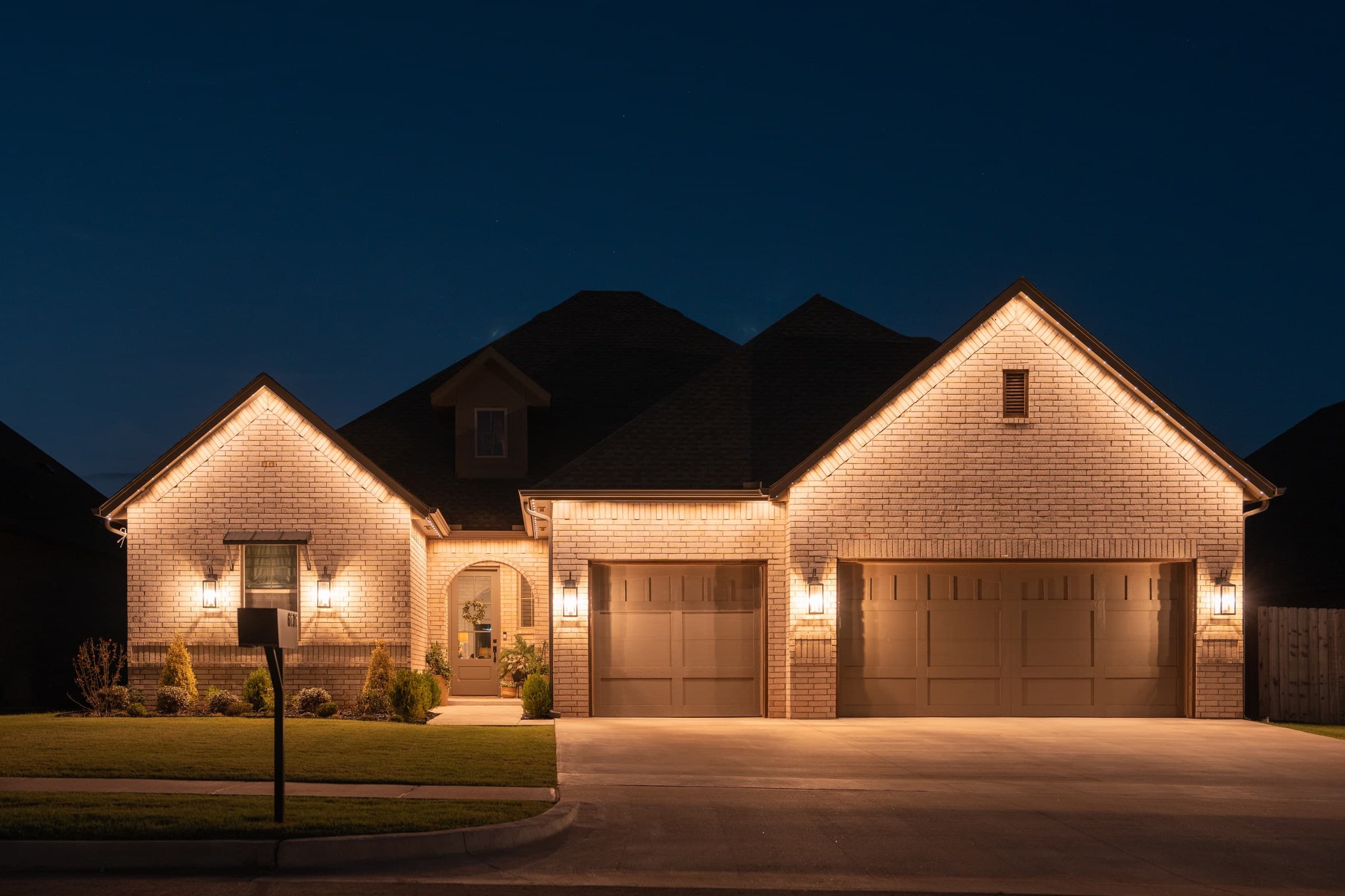 A two-gable brick home at night lit entirely in warm white, with the light continuing down the garage elevation