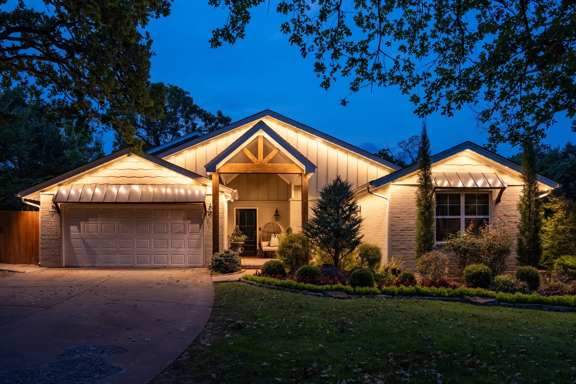 A timber-entry home at blue hour under mature trees, roofline and gable peaks picked out in warm white